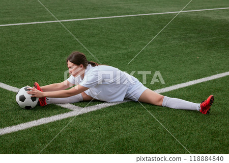 A young female soccer player stretches on a soccer field and reaches for a soccer ball A young female soccer player stretches on a soccer field and reaches for a soccer ball 118884840