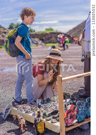 Mother and son tourists buying Mexican souvenirs from a street vendor at the foot of the Teotihuacan pyramid. Cultural experience, travel, and local craftsmanship concept 118885032