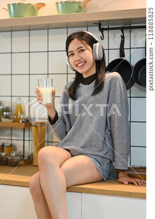 Cheerful young woman sitting in a kitchen with a glass of milk and enjoying music on headphones 118885825