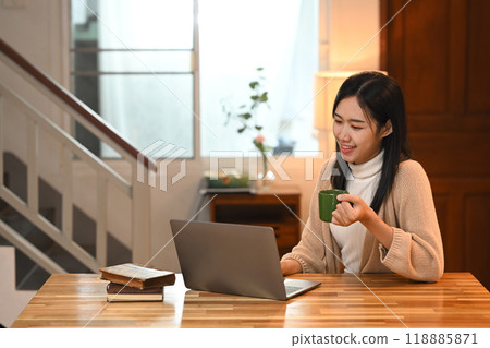 Happy young asian woman drinking hot coffee and using laptop on kitchen counter 118885871