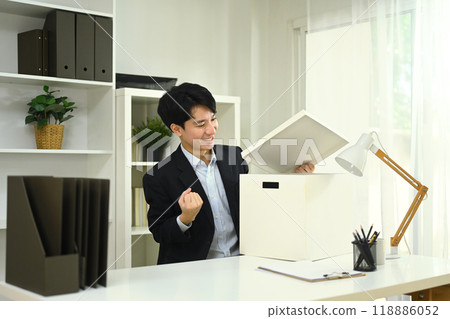Young businessman sitting at office desk, opening a storage box with an excited and satisfied expression Young businessman sitting at office desk, opening a storage box with an excited and satisfied expression 118886052