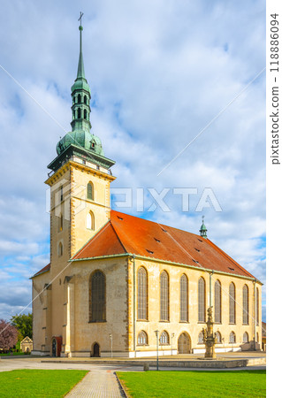 The Church of the Assumption of the Virgin Mary showcases stunning late Gothic architecture in Most, Czechia, with its tall spire and vibrant orange roof against a cloudy sky. 118886094