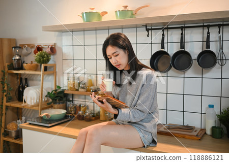 Beautiful young woman sitting on a kitchen countertop, holding a glass of milk and reading a book 118886121