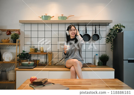 Happy young Asian woman sitting at a kitchen counter with a glass of milk and enjoying music Happy young Asian woman sitting at a kitchen counter with a glass of milk and enjoying music 118886181
