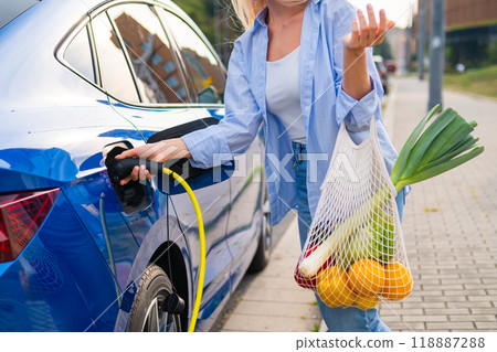A blonde woman plugs in her electric vehicle as she holds a mesh bag filled with fresh vegetables outside. 118887288