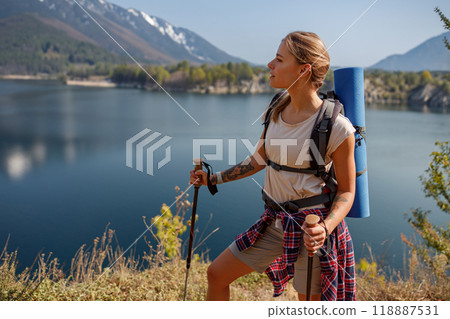 A woman hiker with a backpack enjoys scenic views near a lake and stunning mountains A woman hiker with a backpack enjoys scenic views near a lake and stunning mountains 118887531