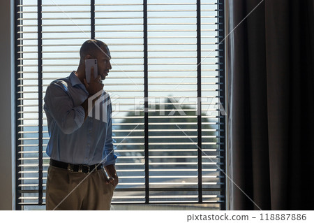 Man wearing shirt posing near big window in office and talking on mobile phone 118887886
