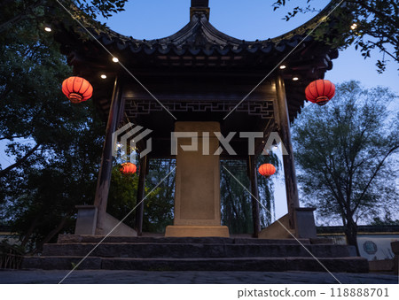A traditional Chinese pavilion illuminated by lanterns during twilight in a serene garden setting with lush trees 118888701