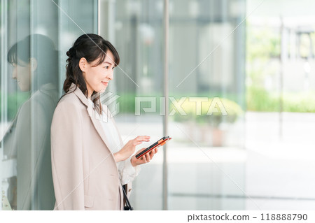 Business women and working mothers waiting to meet while looking at smartphones in front of office buildings and commercial facilities Business women and working mothers waiting to meet while looking at smartphones in front of office buildings and commercial facilities 118888790