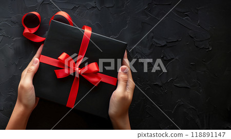 Close-up of hands holding a black gift box with red ribbon on a black background with copy space for text for Black Friday event. AI generated. 118891147