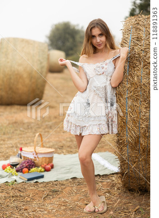 Sexy woman in white dress having picnic standing by hay bales in farm field 118891363
