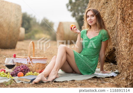 Young beautiful sexy woman in summer green dress sitting and eating ripe apple on soft mat near stack of straw, romantic picnic in field 118891514