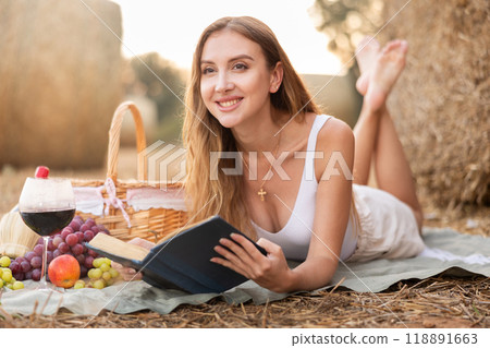 Beautiful woman in white blouse and shorts lying down and and reading book near haystacks with basket of fruit and wine, fashion concept Beautiful woman in white blouse and shorts lying down and and reading book near haystacks with basket of fruit and wine, fashion concept 118891663