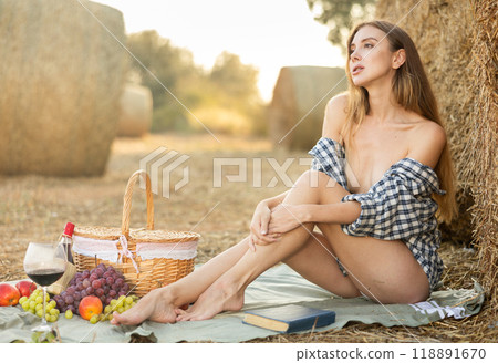 Beautiful young woman in checkered shirt resting near haystack in middle of wheat field on summer evening 118891670