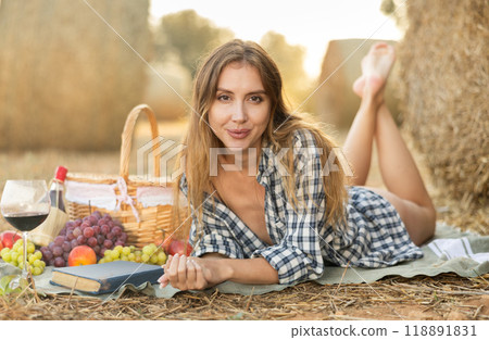 Beautiful cowboy woman in checked shirt lying down and posing near haystacks with basket of fruit and wine, fashion concept 118891831