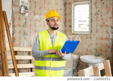 An architect in protective hard hat and yellow vest checks room renovation plan using laptop 118891852
