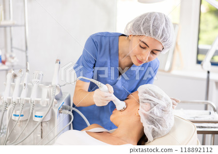 Young female cosmetologist doing ultrasound procedure to young woman 118892112