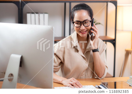 Young asian business woman using desktop computer while talking with smart phone in home office. 118892385