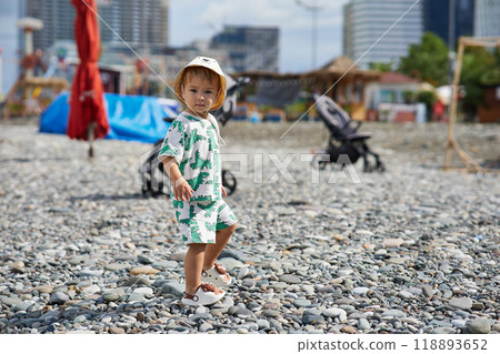 A toddler in a cute outfit and a sunhat walks along a rocky beach near the ocean as waves crash nearby. The scene captures the child's adventurous spirit and the natural beauty of the stormy  118893652