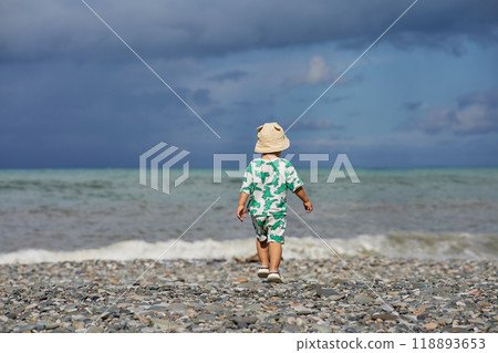 A toddler in a cute outfit and a sunhat walks along a rocky beach near the ocean as waves crash nearby. The scene captures the child's adventurous spirit and the natural beauty of the stormy  118893653