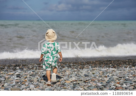 A toddler in a cute outfit and a sunhat walks along a rocky beach near the ocean as waves crash nearby. The scene captures the child's adventurous spirit and the natural beauty of the stormy  118893654