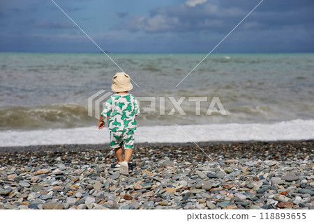 A toddler in a cute outfit and a sunhat walks along a rocky beach near the ocean as waves crash nearby. The scene captures the child's adventurous spirit and the natural beauty of the stormy A toddler in a cute outfit and a sunhat walks along a rocky beach near the ocean as waves crash nearby. The scene captures the child's adventurous spirit and the natural beauty of the stormy 118893655