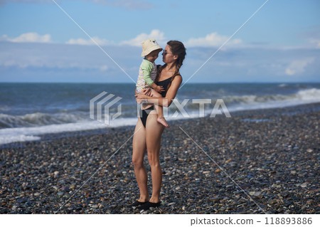 A young mother in sunglasses holds her toddler while standing on a pebble beach by the ocean. The child wears a sunhat and swimsuit, and the waves crash behind them, capturing a joyful family moment. A young mother in sunglasses holds her toddler while standing on a pebble beach by the ocean. The child wears a sunhat and swimsuit, and the waves crash behind them, capturing a joyful family moment. 118893886