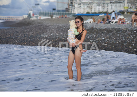 A young mother in sunglasses holds her toddler while standing on a pebble beach by the ocean. The child wears a sunhat and swimsuit, and the waves crash behind them, capturing a joyful family moment. 118893887
