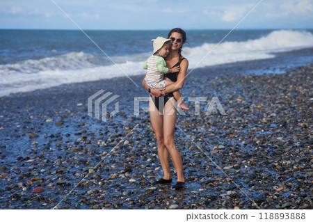 A young mother in sunglasses holds her toddler while standing on a pebble beach by the ocean. The child wears a sunhat and swimsuit, and the waves crash behind them, capturing a joyful family moment. A young mother in sunglasses holds her toddler while standing on a pebble beach by the ocean. The child wears a sunhat and swimsuit, and the waves crash behind them, capturing a joyful family moment. 118893888