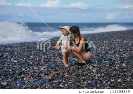 A young mother in sunglasses holds her toddler while standing on a pebble beach by the ocean. The child wears a sunhat and swimsuit, and the waves crash behind them, capturing a joyful family moment. A young mother in sunglasses holds her toddler while standing on a pebble beach by the ocean. The child wears a sunhat and swimsuit, and the waves crash behind them, capturing a joyful family moment. 118893889