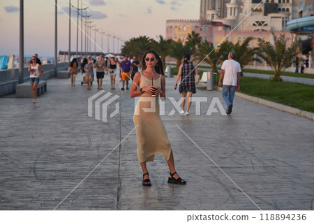 A stylish woman in a light summer dress walks confidently along a busy promenade at sunset, holding a cup of coffee. The background features palm trees, people, and seaside buildings, creating a relax 118894236