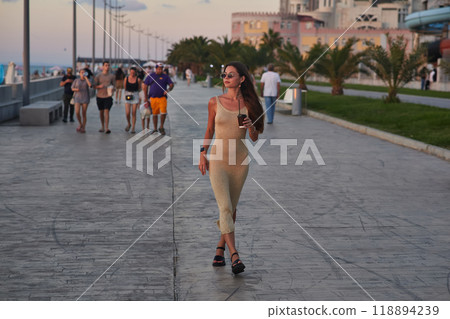A stylish woman in a light summer dress walks confidently along a busy promenade at sunset, holding a cup of coffee. The background features palm trees, people, and seaside buildings, creating a relax 118894239