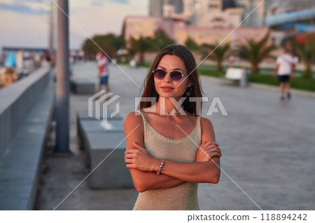 A stylish woman in a light summer dress walks confidently along a busy promenade at sunset, holding a cup of coffee. The background features palm trees, people, and seaside buildings, creating a relax A stylish woman in a light summer dress walks confidently along a busy promenade at sunset, holding a cup of coffee. The background features palm trees, people, and seaside buildings, creating a relax 118894242