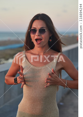 A stylish woman in a light summer dress walks confidently along a busy promenade at sunset, holding a cup of coffee. The background features palm trees, people, and seaside buildings, creating a relax 118894244