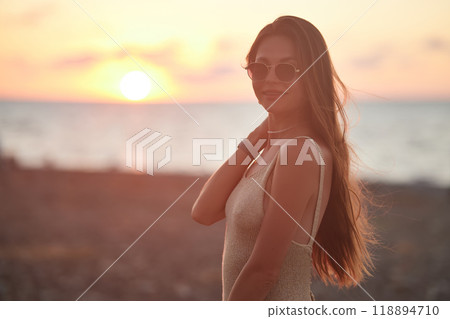 A serene sunset moment captured by the sea. A young woman, with long hair and wearing sunglasses, stands in profile against the glowing horizon. She radiates calmness in the golden hour. 118894710