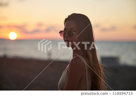 A serene sunset moment captured by the sea. A young woman, with long hair and wearing sunglasses, stands in profile against the glowing horizon. She radiates calmness in the golden hour. 118894713