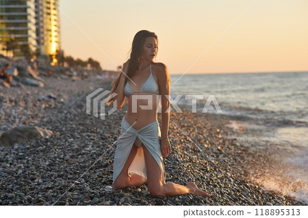 A young woman in a bikini kneels on a rocky beach at sunset. The soft evening light highlights her hair and silhouette as gentle waves wash up on the shore. Peaceful moment by the sea. 118895313