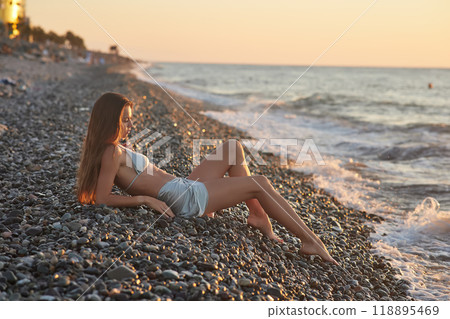 A woman relaxes on a rocky beach at sunset, wearing a bikini and cover-up skirt. The warm golden light of the setting sun reflects on the ocean, creating a serene, peaceful, and scenic atmosphere. 118895469