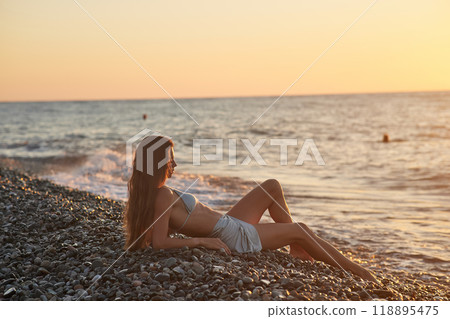 A woman relaxes on a rocky beach at sunset, wearing a bikini and cover-up skirt. The warm golden light of the setting sun reflects on the ocean, creating a serene, peaceful, and scenic atmosphere. A woman relaxes on a rocky beach at sunset, wearing a bikini and cover-up skirt. The warm golden light of the setting sun reflects on the ocean, creating a serene, peaceful, and scenic atmosphere. 118895475