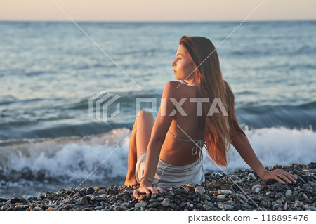 A woman relaxes on a rocky beach at sunset, wearing a bikini and cover-up skirt. The warm golden light of the setting sun reflects on the ocean, creating a serene, peaceful, and scenic atmosphere. 118895476