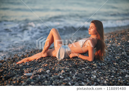 A woman relaxes on a rocky beach at sunset, wearing a bikini and cover-up skirt. The warm golden light of the setting sun reflects on the ocean, creating a serene, peaceful, and scenic atmosphere. A woman relaxes on a rocky beach at sunset, wearing a bikini and cover-up skirt. The warm golden light of the setting sun reflects on the ocean, creating a serene, peaceful, and scenic atmosphere. 118895480