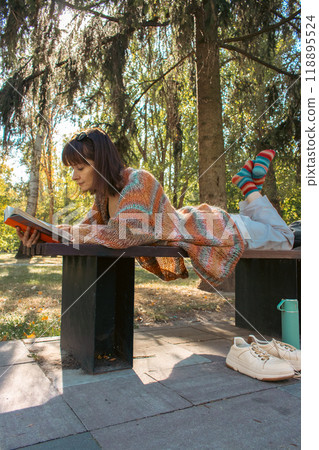 Brunette woman lying on bench with open book. Student reading book in the park. Girl in stylish knitted sweater studying in campus park. Education concept. Student lifestyle. Read and relax. 118895524
