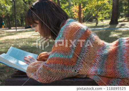 Brunette woman lying on bench with open book. Student reading book in the park. Girl in stylish knitted sweater studying in campus park. Education concept. Student lifestyle. Read and relax. 118895525