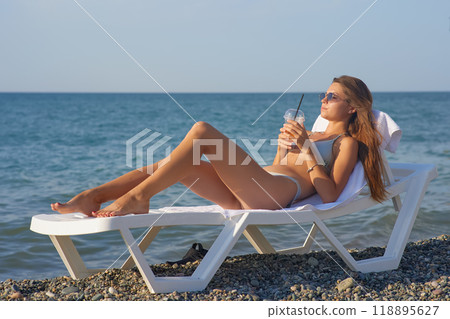 A young woman relaxes on a beach lounge chair by the ocean, sipping a refreshing drink under the warm sun. The serene coastal scenery and clear blue water capture the essence of a perfect summer day. A young woman relaxes on a beach lounge chair by the ocean, sipping a refreshing drink under the warm sun. The serene coastal scenery and clear blue water capture the essence of a perfect summer day. 118895627