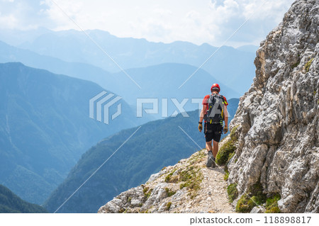 A hiker navigates a narrow trail high in the Julian Alps, surrounded by stunning mountain vistas and lush green valleys under a partly cloudy sky. 118898817