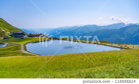 This picturesque view features the Wildkogel water reservoir surrounded by lush green hills in the Austrian Alps. Visitors enjoy the scenic beauty in bright, clear weather. This picturesque view features the Wildkogel water reservoir surrounded by lush green hills in the Austrian Alps. Visitors enjoy the scenic beauty in bright, clear weather. 118898824