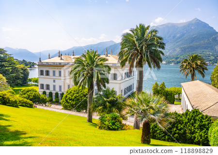 Nestled by the tranquil waters of Lake Como, Villa Melzi showcases neoclassical architecture amidst lush gardens and majestic mountains under a bright blue sky. Nestled by the tranquil waters of Lake Como, Villa Melzi showcases neoclassical architecture amidst lush gardens and majestic mountains under a bright blue sky. 118898829