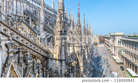 A stunning view from the rooftop of the Duomo in Milan, showcasing intricate gothic details and the bustling city streets below under a clear blue sky. A stunning view from the rooftop of the Duomo in Milan, showcasing intricate gothic details and the bustling city streets below under a clear blue sky. 118898841
