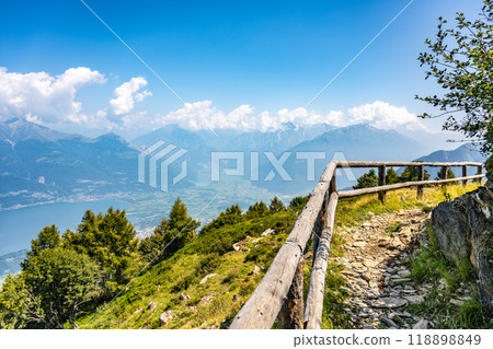 A breathtaking panorama reveals Lake Como nestled between lush hills, framed by majestic mountains and clear blue skies. Nature's beauty captivates visitors exploring the scenic trails. 118898849
