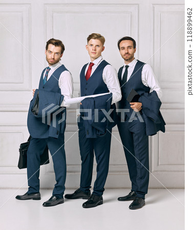 Discussing business after productive day. Young men dressed formal attire posing, against white background with blazers in hands. Discussing business after productive day. Young men dressed formal attire posing, against white background with blazers in hands. 118899462